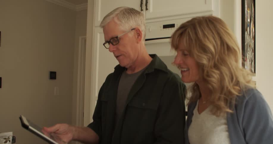 A woman walks into her kitchen and stops to look at a tablet with her husband