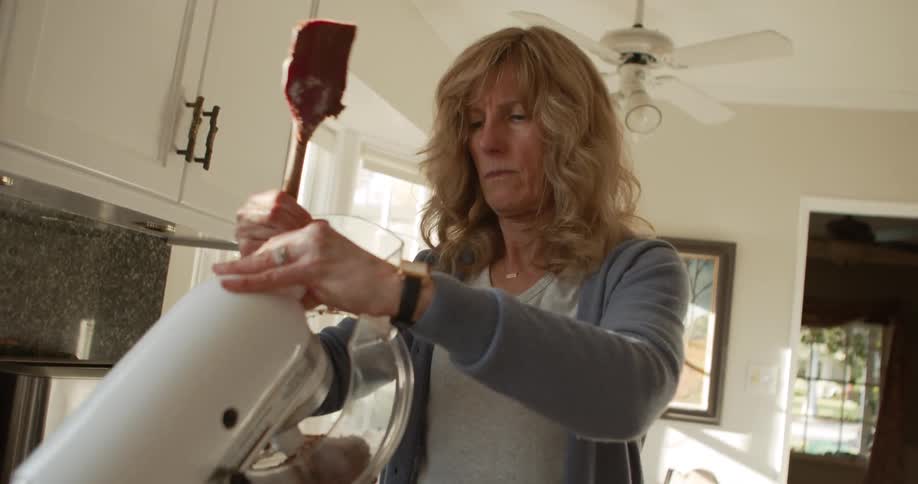 A woman stops a standing mixer and lifts the top to clean the blades with a spatula