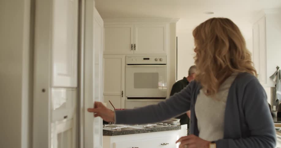 A woman goes to the fridge and a man goes to a cupboard as they cook together in their kitchen