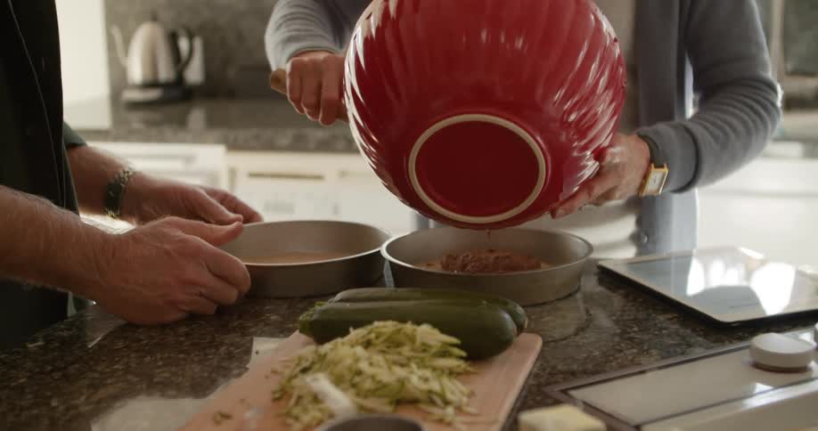 A woman pours cake batter in to a pan and a man slides another pan to her