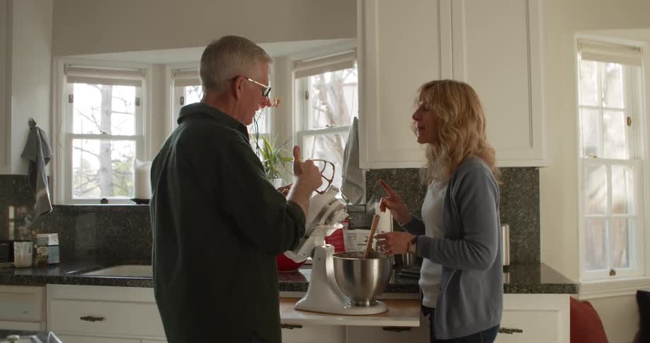 Man wearing glasses a dark shirt and blonde woman use a stand mixer and taste food in the kitchen
