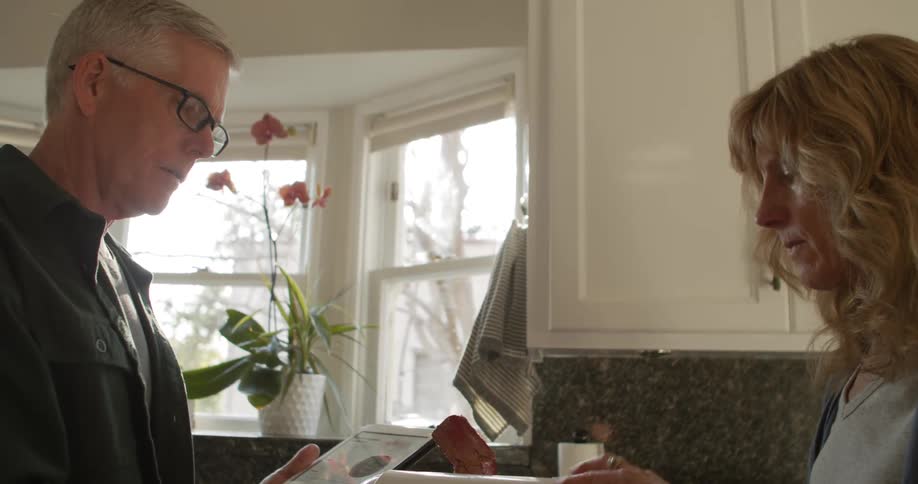 A woman uses a blender while speaking with a man using a tablet standing across from her in a kitchen during the day