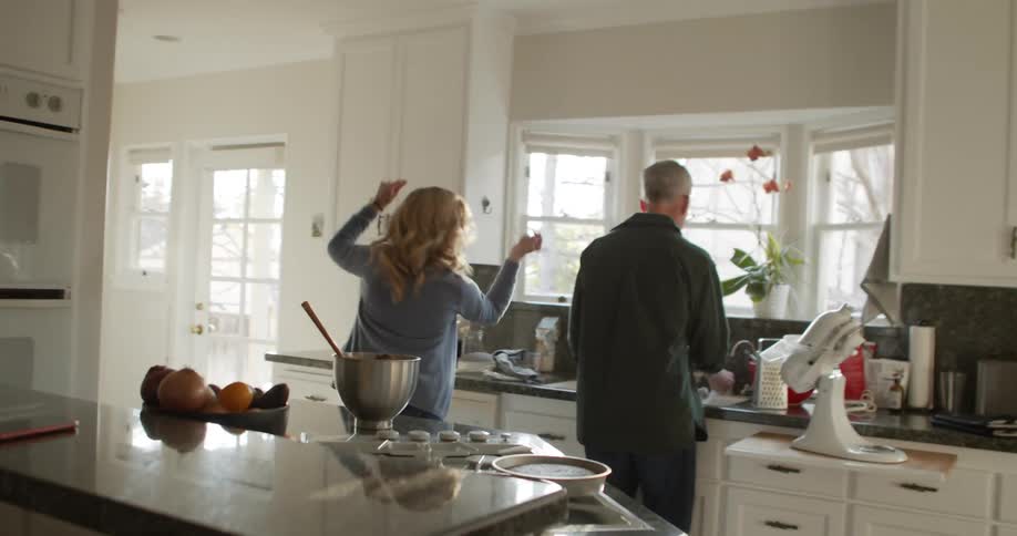 A woman snaps her fingers as she twirls and dances next to her husband at the kitchen sink