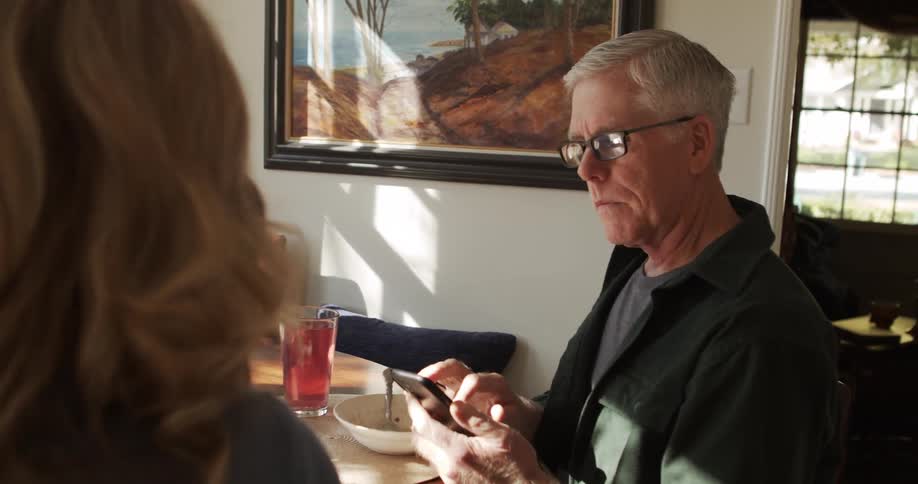 A man and woman sit at a table at home eating while he uses a cell phone and they talk