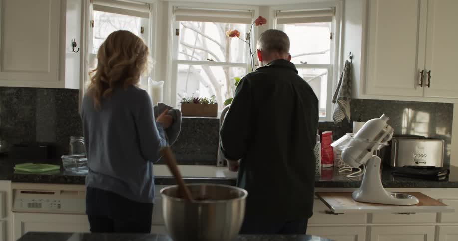 A man and woman talk and smile as they wash and dry dishes together in their sunny white kitchen