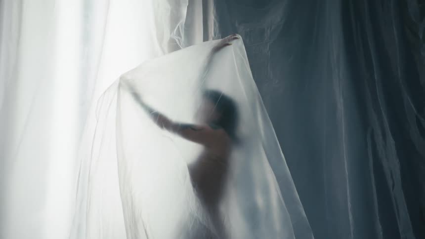 Standing nude woman with tattoos moves under gray filmy fabric