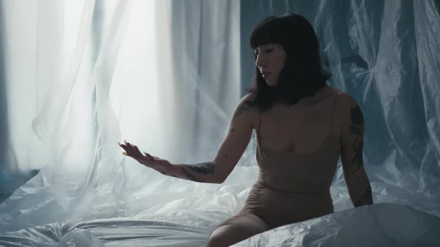 A woman sits on a bed covered in filmy plastic and with plastic sheets hanging looking at her hand