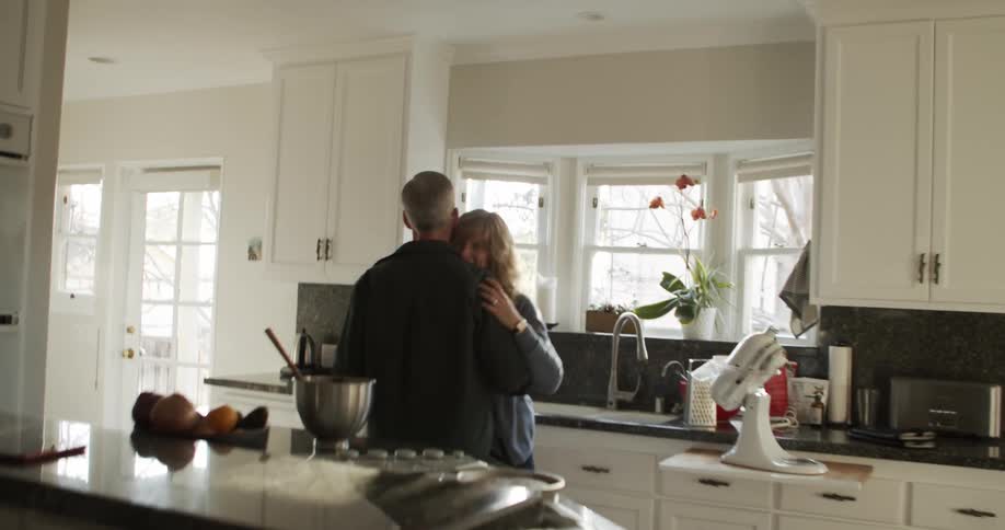 Husband with glasses and a dark shirt and blonde wife in a blue sweater dance in the kitchen