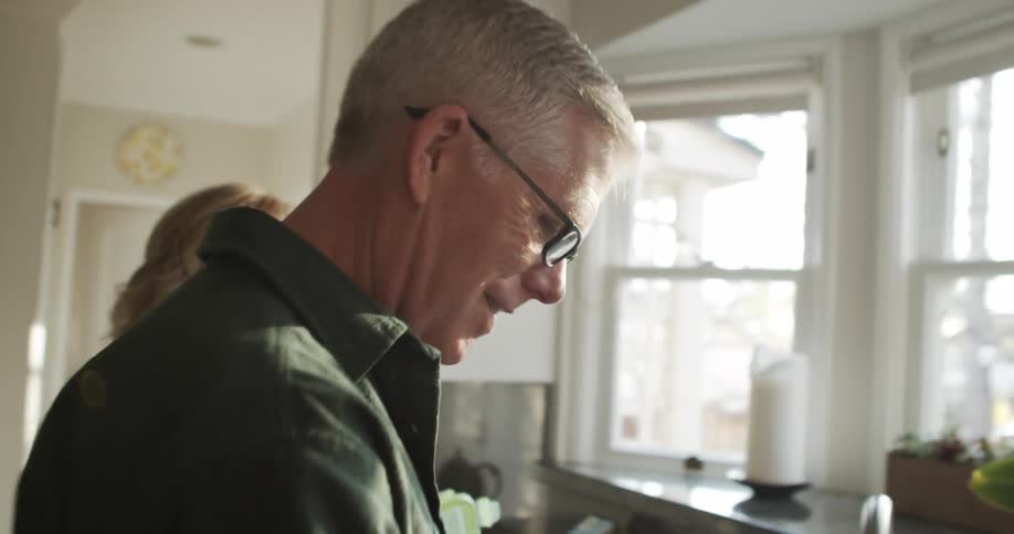 Man wearing glasses and a dark shirt does the dishes next to blonde woman in the kitchen
