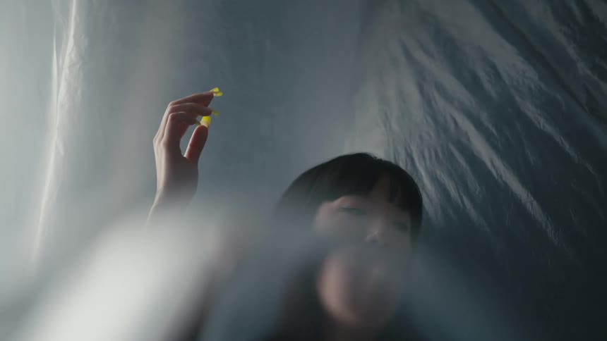 Woman with yellow polish tipped long fingernails raises hand against backdrop of filmy gray fabric