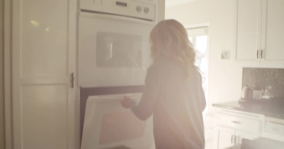 A woman in a home kitchen puts on an oven mitt and opens an oven to remove the food cooking in a pan within