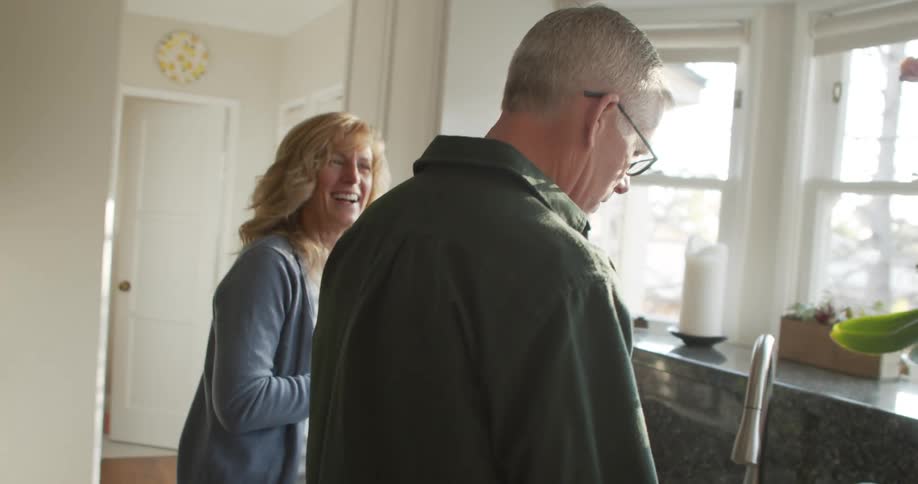 A woman laughs and speaks with a man as she carries food containers through a home kitchen during the day