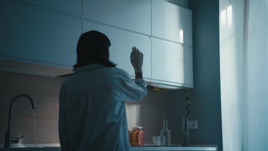 A woman stands in a kitchen with the faucet running and holds her hand in front of her face and examines it