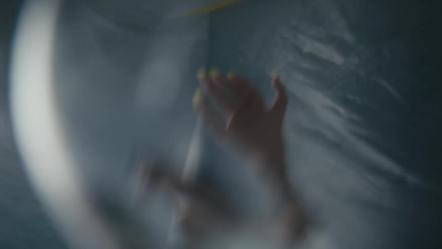 Woman with long yellow fingernails can be seen through glass reaching upward near silver gray fabric