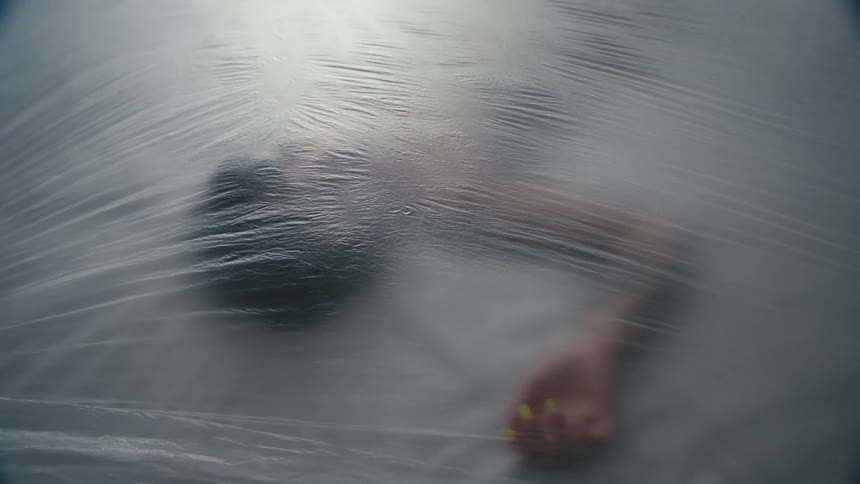 A plastic sheet falls over a woman lying motionlessly on bed covered in plastic
