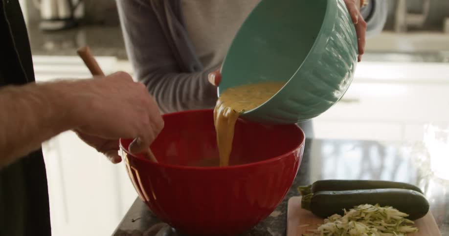 Couple cook together adding mixture to another bowl near wooden chopping board holding whole and shredded zucchini