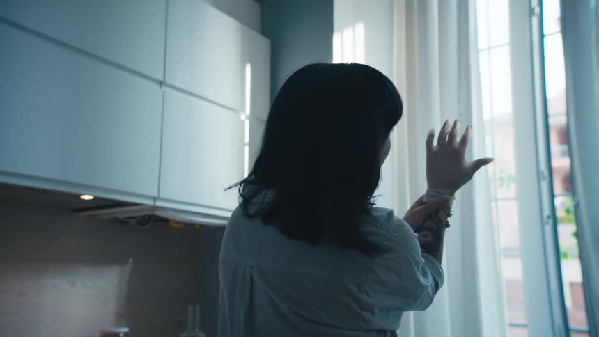 Woman wearing gloves with long fingernails stands in kitchen near window examining hand