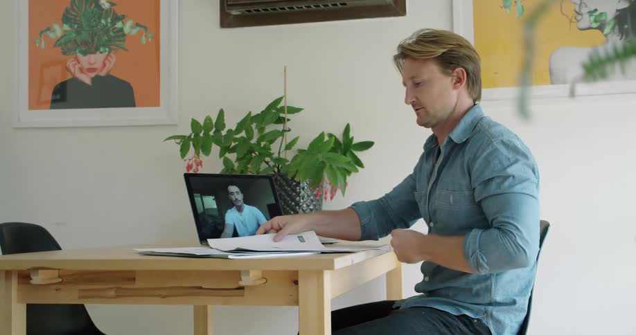 A man sits at a desk and video chats with a man on a laptop