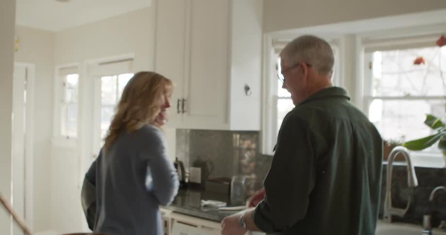 A woman playfully whips her husband with a washcloth causing him to stop washing the dishes and snatch away the washcloth before kissing her