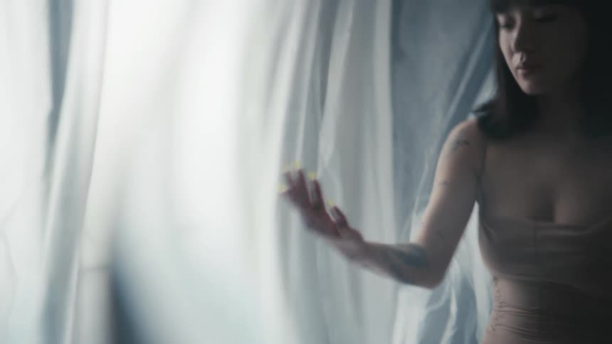 A woman sits in a room draped in sheer fabric as she stares at her own hand