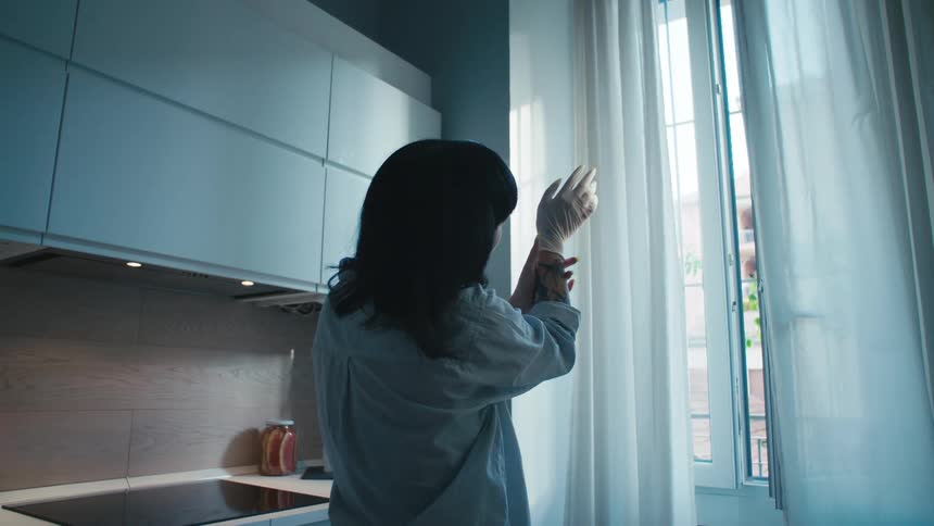 A woman stands in front of a window in her kitchen and holds her hand up and examines it