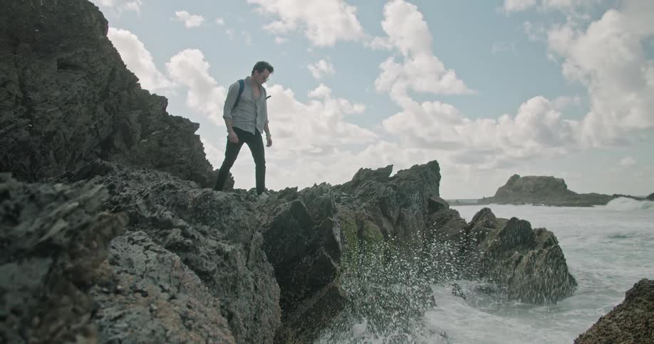 Man with a grey shirt a black backpack and dark pants wanters around rocky shore