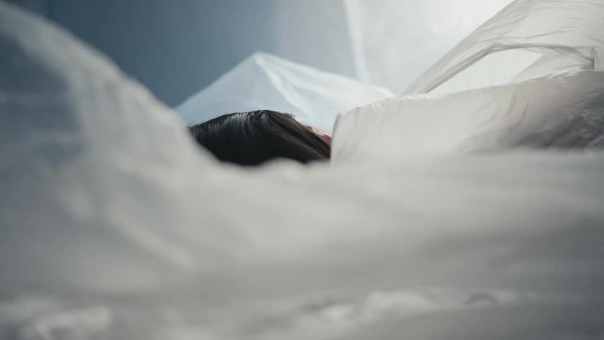 A woman in a room laying on a bed with a plastic sheet being pulled off of her with a wall in the background