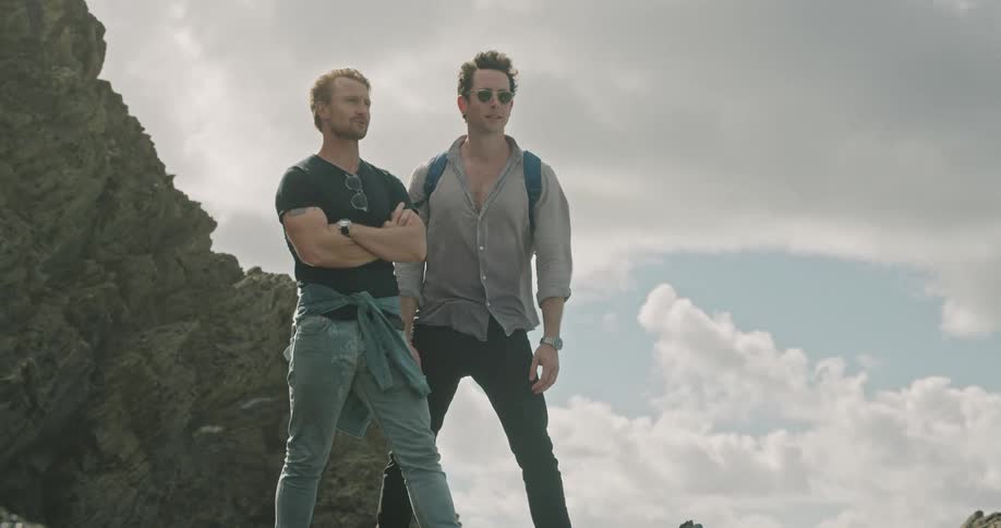 Two men stand on rocky mountainous terrain while one points off into the distance
