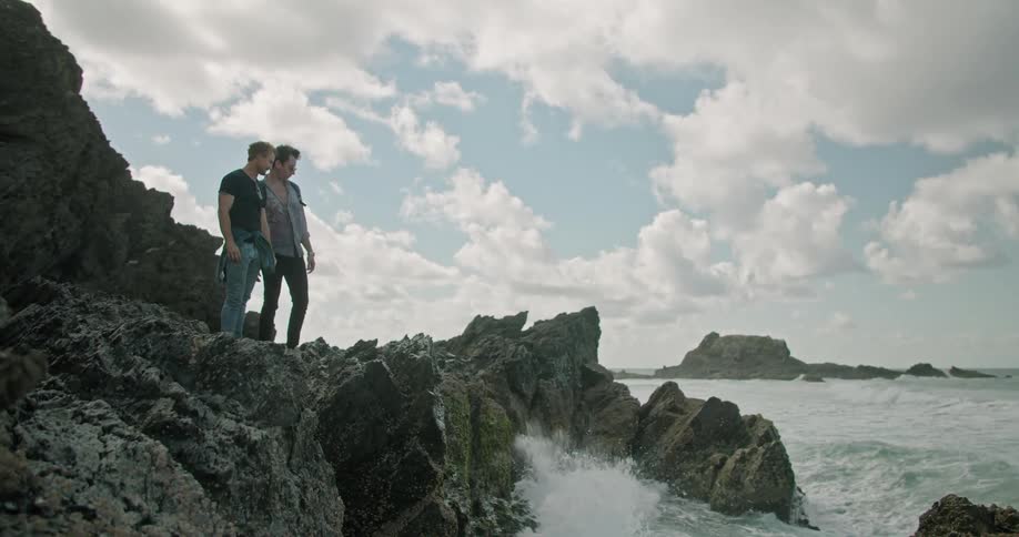Two young men stand on a rocky cliff and watch as waves crash against it