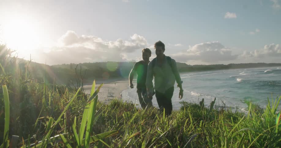 Two men walk up a grassy slope from the beach on a sunny day