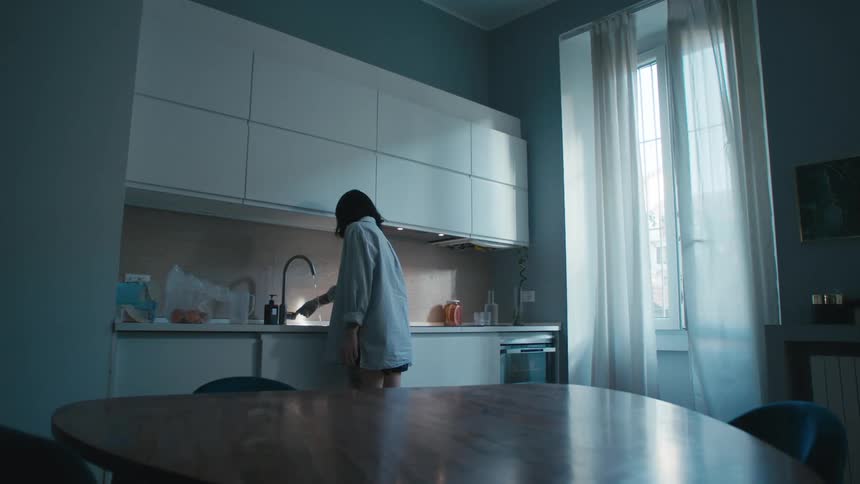A woman stands indoors in front of a sink and turns on the faucet