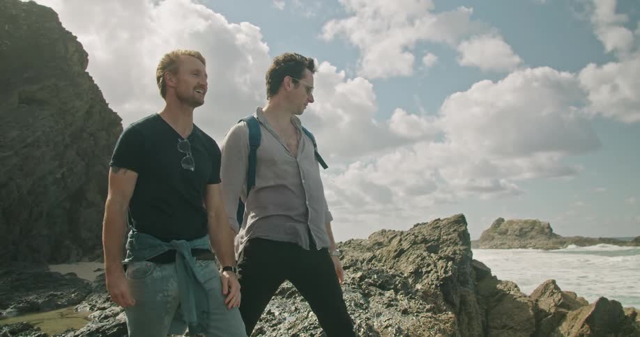 Two men stand on the shore and watch water spray up on rocks and hug and high five one another