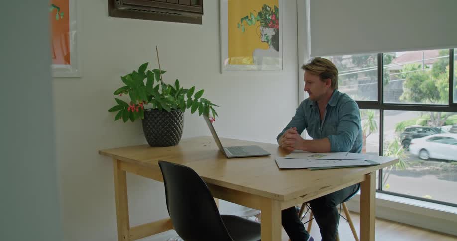 A man sits at a table with a laptop on it and waves to the person he is teleconferencing with