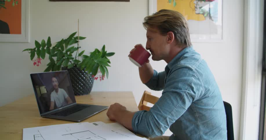 A man drinks coffee and talks to his colleague via video chat on a laptop