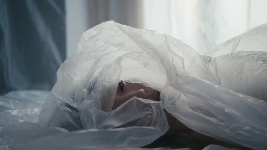 A woman laying in a room covered in plastic with a window and curtain in the background