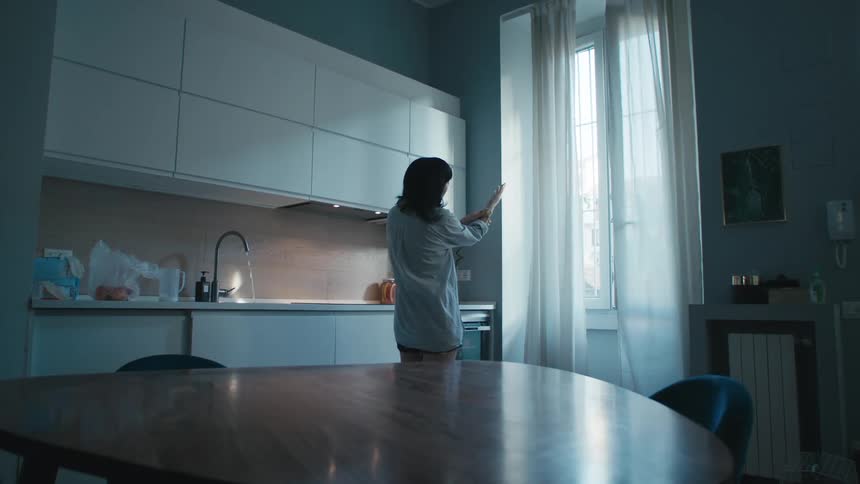 A woman holds up and looks at her hands while standing in front of a window