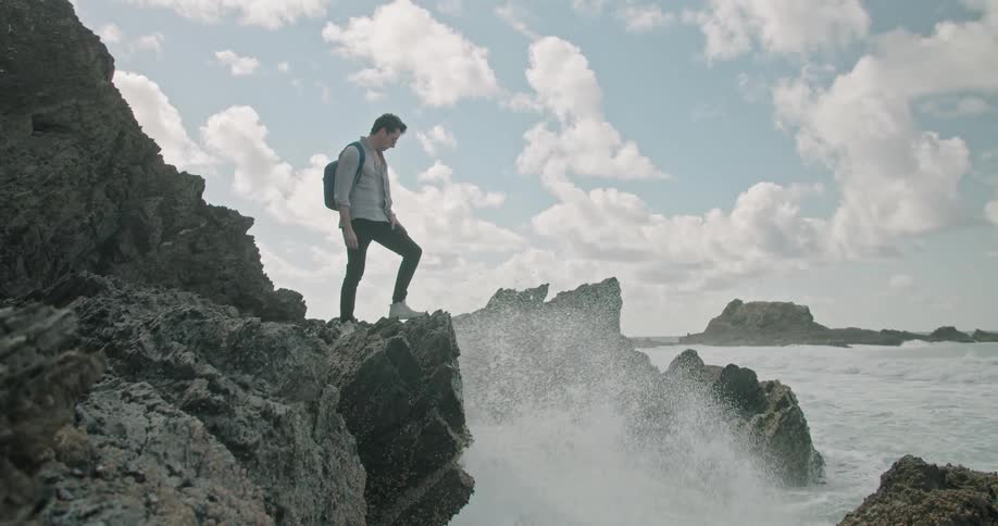 A man looks out over the ocean while standing on a rock formation on the coast during the day