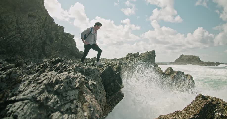 A man hikes across a cliff and leans over and looks at the water on the shore splashing up on the rocks