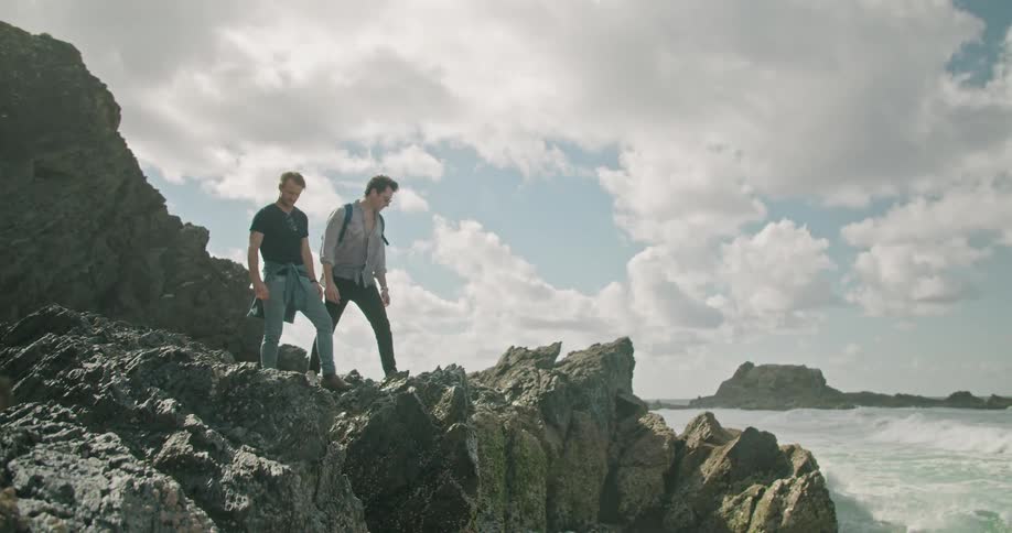 A pair of men standing on a rock formation on the coast lean forward to gaze over the edge at the ocean below on a sunny day