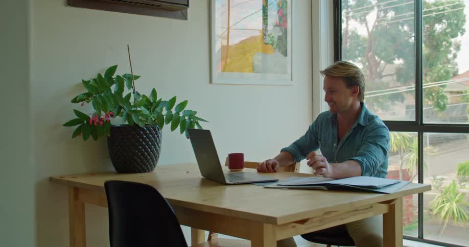 A man waves and smiles at the laptop screen as he meets with someone via video call in his office