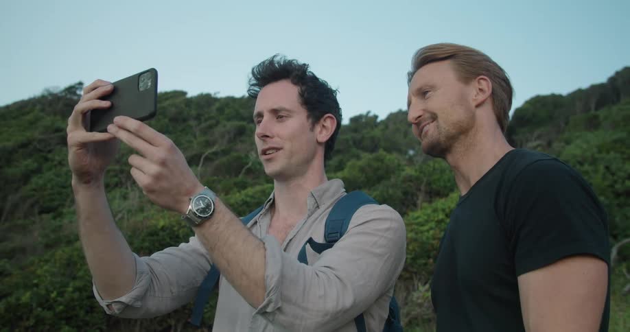 Two men hiking in a forest talk and one takes a picture with his cell phone