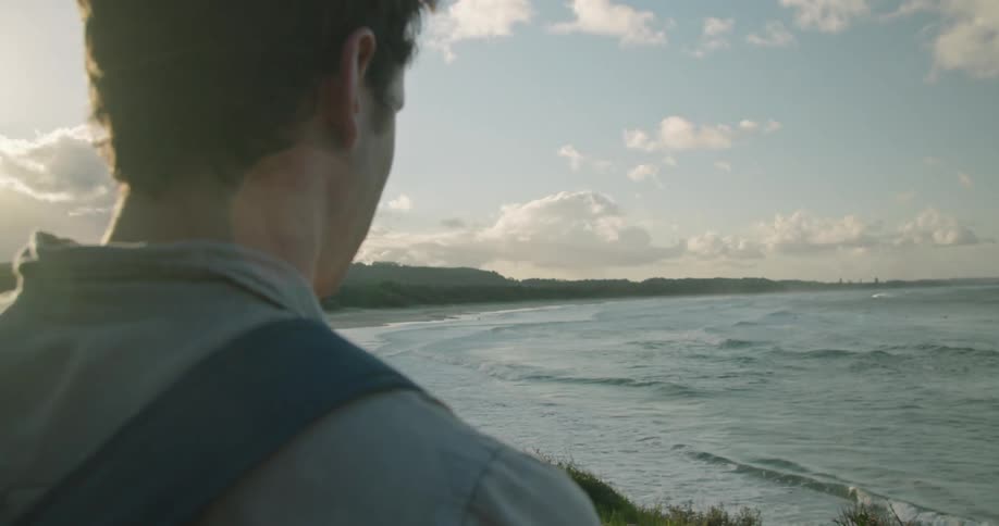 A man standing on a hill over a beach uses his smartphone to take a picture of the ocean before turning and walking out of frame