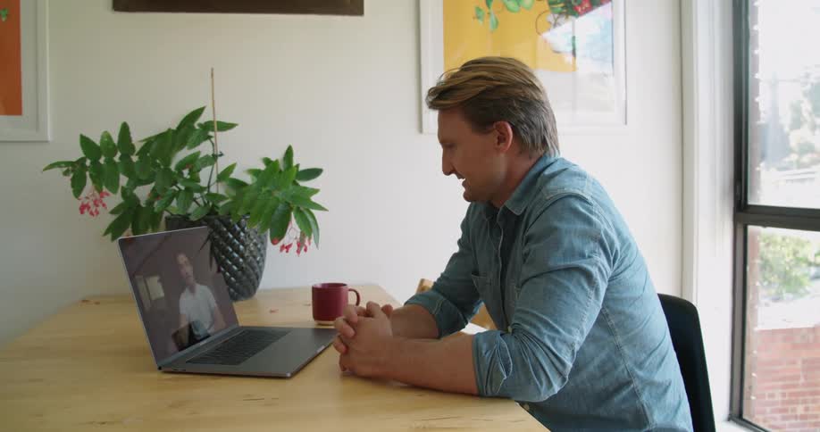 A man in a room sitting on a chair at a table using a laptop