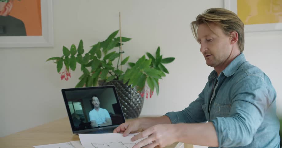 A man nods and picks up plans as he talks and meets with another on his laptop screen