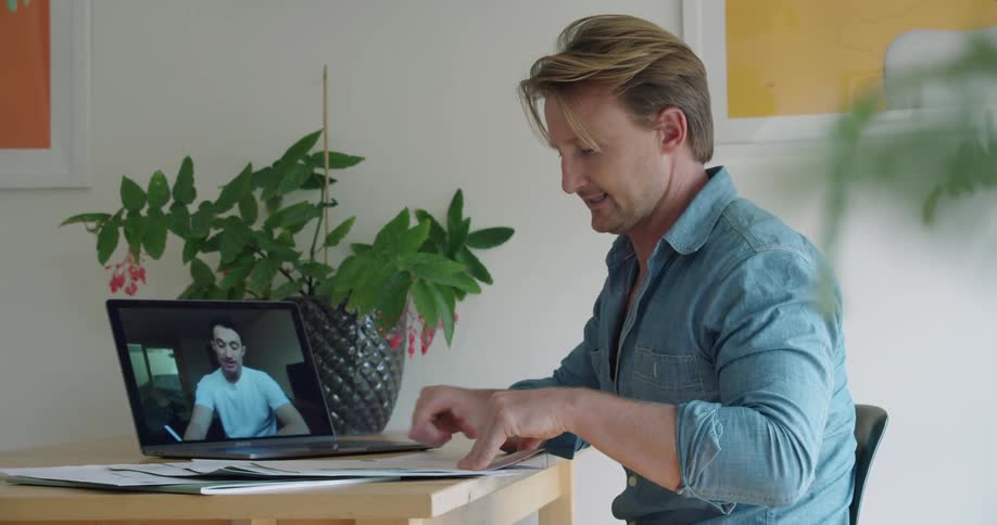 A man sits at a desk with his laptop and some documents during a video conference
