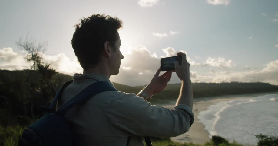 A man stands on a cliff overlooking the ocean and uses his phone to take a photo