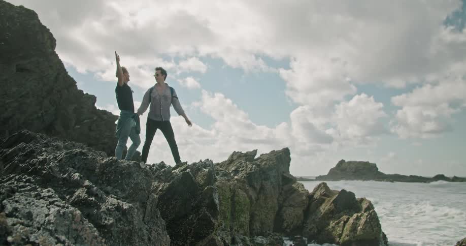 Two men raise up their arms as they stand on the edge of a cliff overlooking the ocean