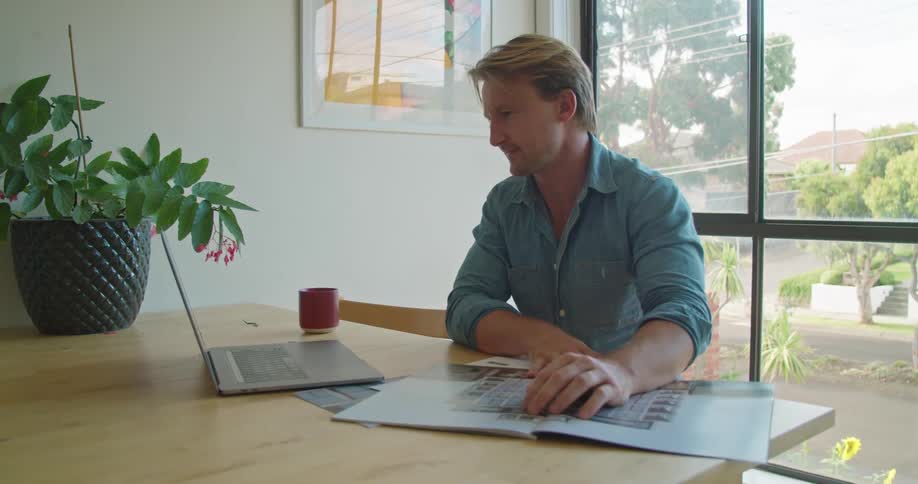 A man opens a book while participating in a video chat on a laptop while sitting at a table indoors during the day
