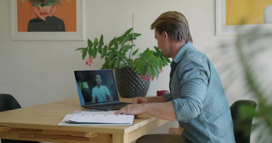 A man searches a stack of floor plans while using a laptop to participate in a video conference at a desk during the day