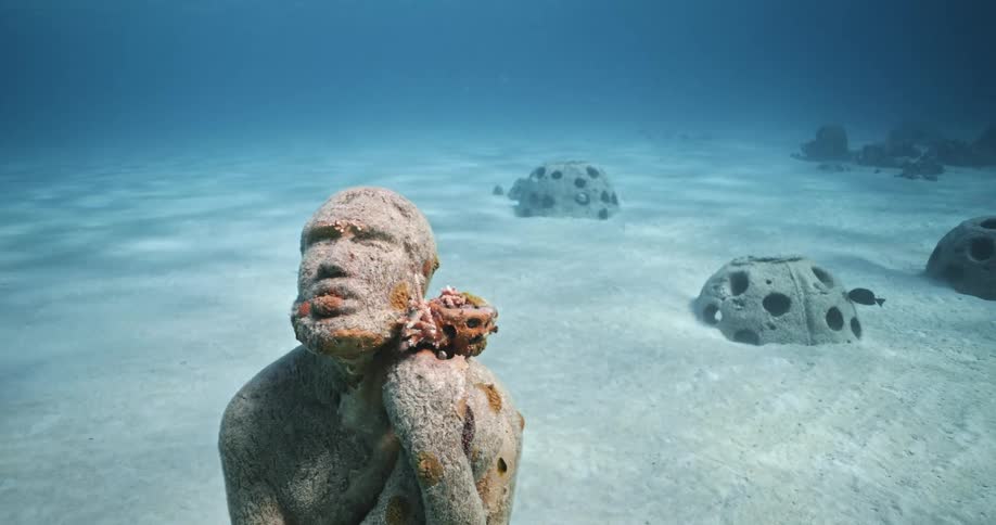 Close up of a statue submerged underwater with fish swimming around in the clear blue ocean water